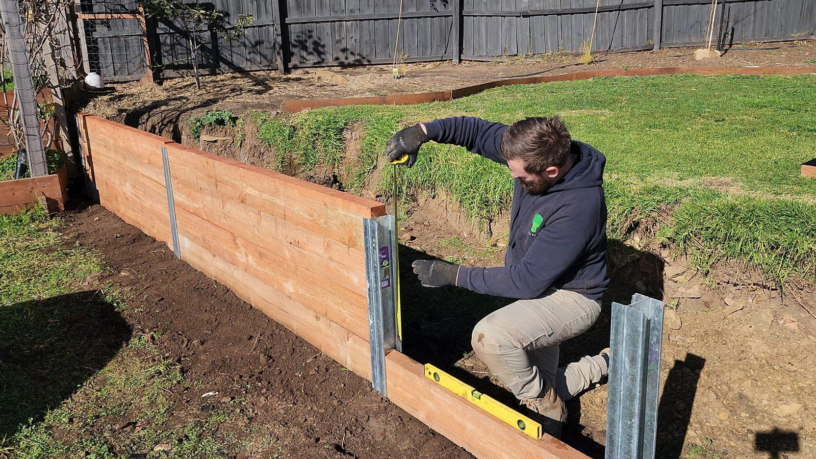 Installer measuring galvanised steel posts while building a timber sleeper retaining wall